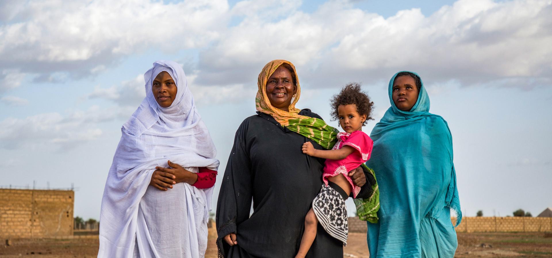Three women stand in long robes, one holding a baby