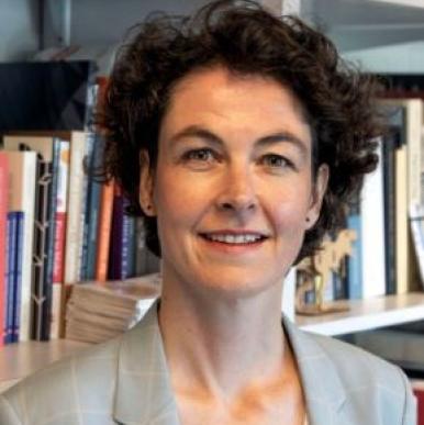 Caucasian woman with short hair in a grey blazer, stnads in front of a bookshelf and looks into the camera