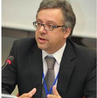 Caucasian man wearing glasses shone speaking into microphone, wearing a black suit