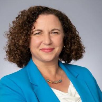 Woman with short, brown curly hair wearing a bright, light blue blazer, looks into the camera