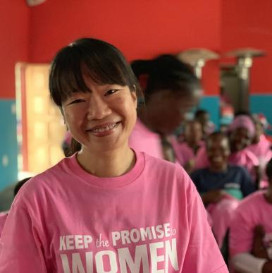 Asian woman with her hair pulled back, wearing a light pink shirt with the words Keep the PROMISE to WOMEN, stands in front of a group of African women and children in the same pink shirts