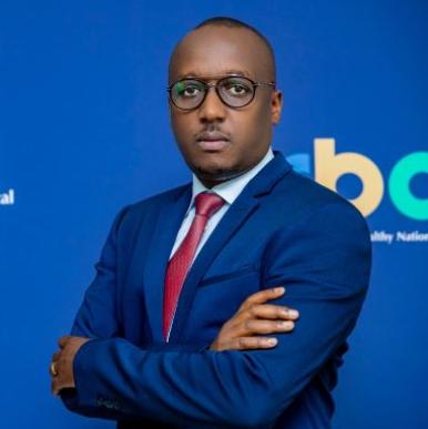 Rwandan man wearing glasses, stands in a bright blue suite with a red tie, against a backdrop that says RBC