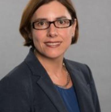 Caucasian woman with short brown hair, wearing glasses and a black blazer, looks into the camera
