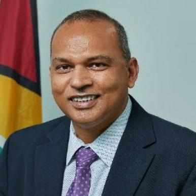 Man in a blue suit with a purple tie sits in front of the flag of Guyana