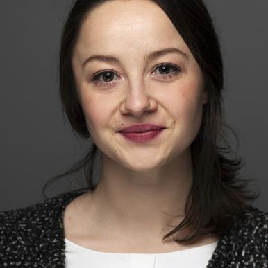 Woman with dark rbown hair, wearing a white blouse and textured jacket,stares into the camera