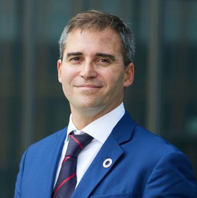 Man in a blue suit, with an SDG pin, stands and looks into camera