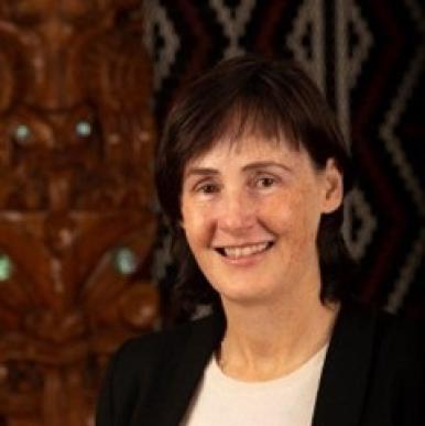 Woman with brown hair with bangs, wearing a white shirt and black blazer, stands and smiles into the camera