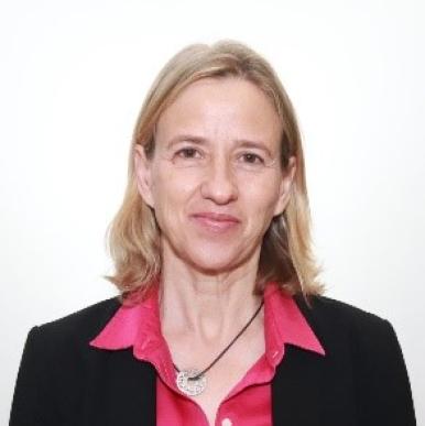 Woman with blonde hair, wearing a pink blouse and black blazer, stands in front of a white background 