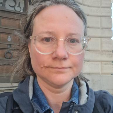 White woman wearing clear glasses and a blue winter jacket looks into the camera