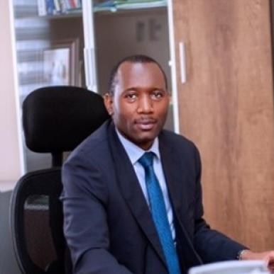 Man wearing a black suit and bright blue tie sits in a chair looks into a camera