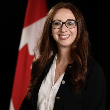 Woman wearing a black blazer and glasses stands in front of a Canadian flag