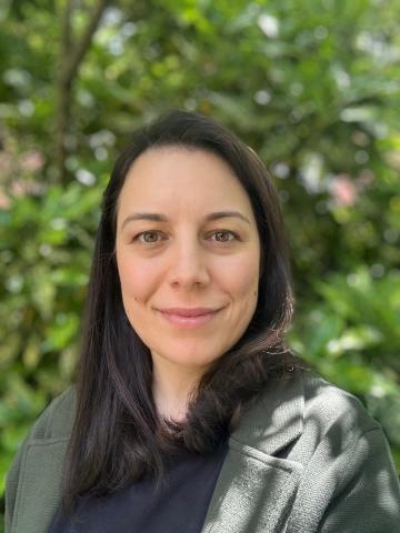 Woman with long brown hair smiles into the camera, standing in front of a green leafy tree