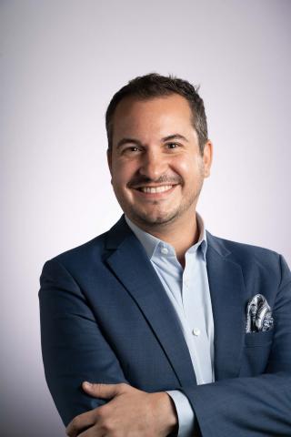 Man wearing navy blue suit with a light blue shirt underneath, crosses his arms and smiles bright into the camera