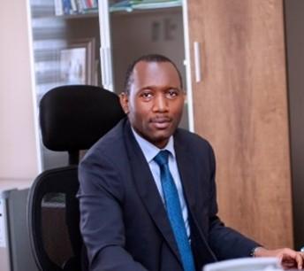 Man wearing a black suit and bright blue tie sits in a chair looks into a camera