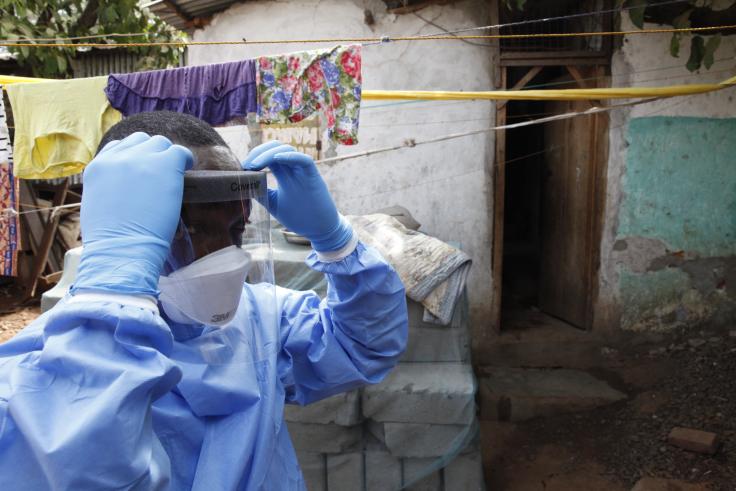 Sierra Leone, PPE Person in full PPE stands outside a home, putting on their face shield