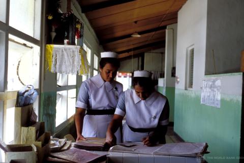 Two women stand in nurses uniforms, learning over and writing into an administrative book